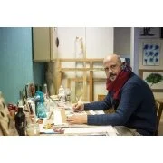 Smiling man seated at a desk with various tools and materials in a workshop or creative space.