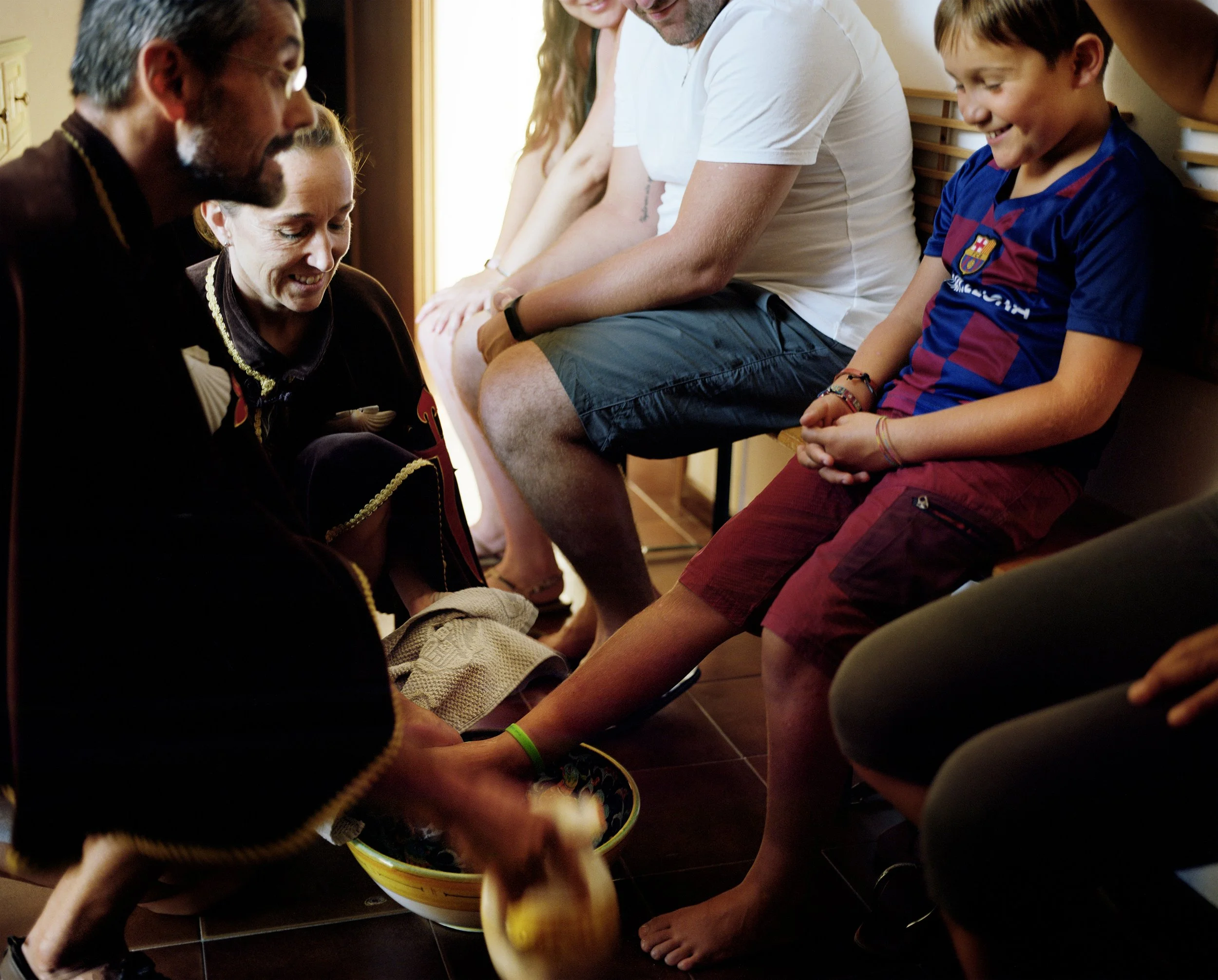 A family seated in a circle, an adult in traditional bearded costume, new to the family, ties a young boy's shoe, the group laughs and shares a convivial moment in a room lit by natural light.