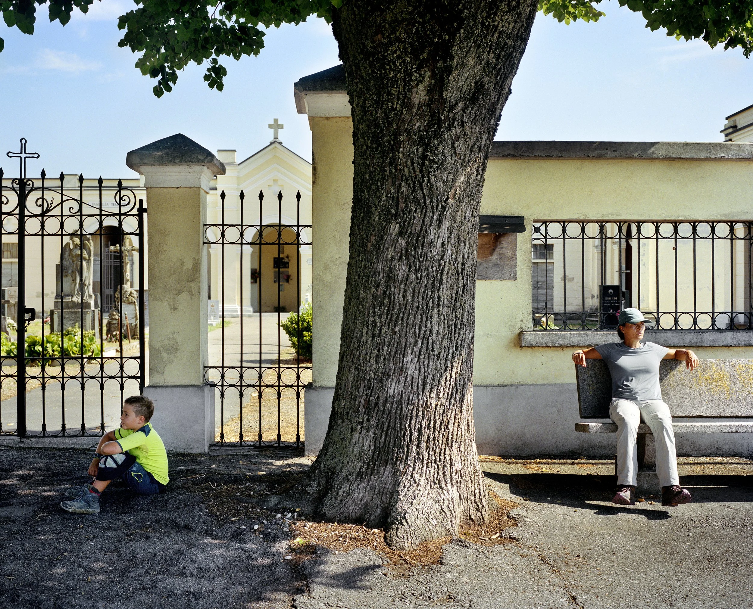 A woman sits on a bench under a large tree, and a young boy sits on the ground beside her, in a mausoleum or cemetery courtyard surrounded by a wrought-iron fence, with graves and funerary statues in the background.