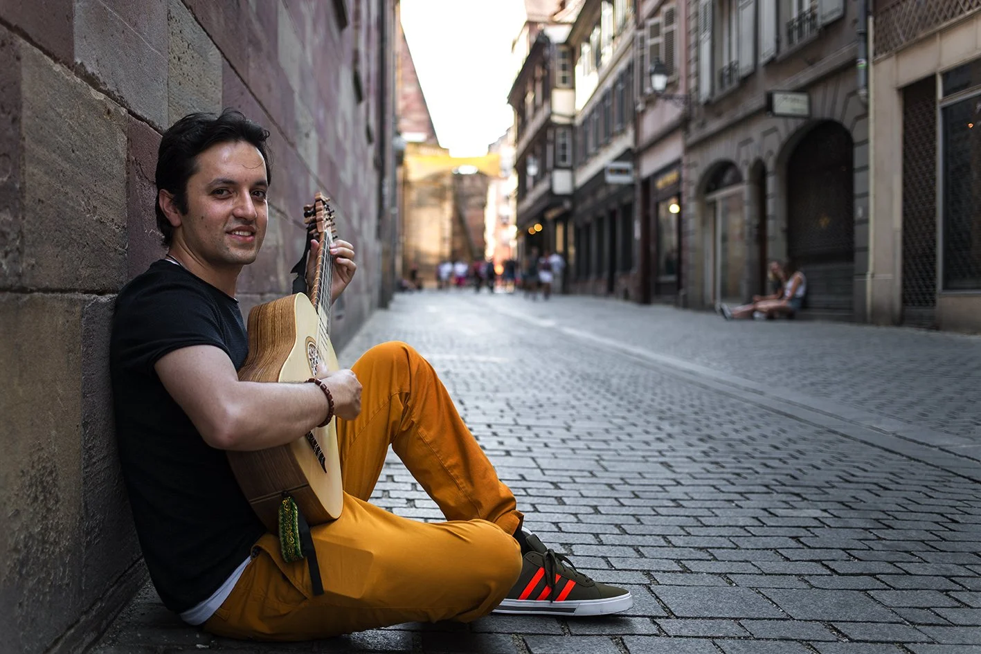 A smiling man sits against a stone wall on a cobbled street, playing his guitar. Other people can be seen in the background, some seated, in an urban atmosphere.