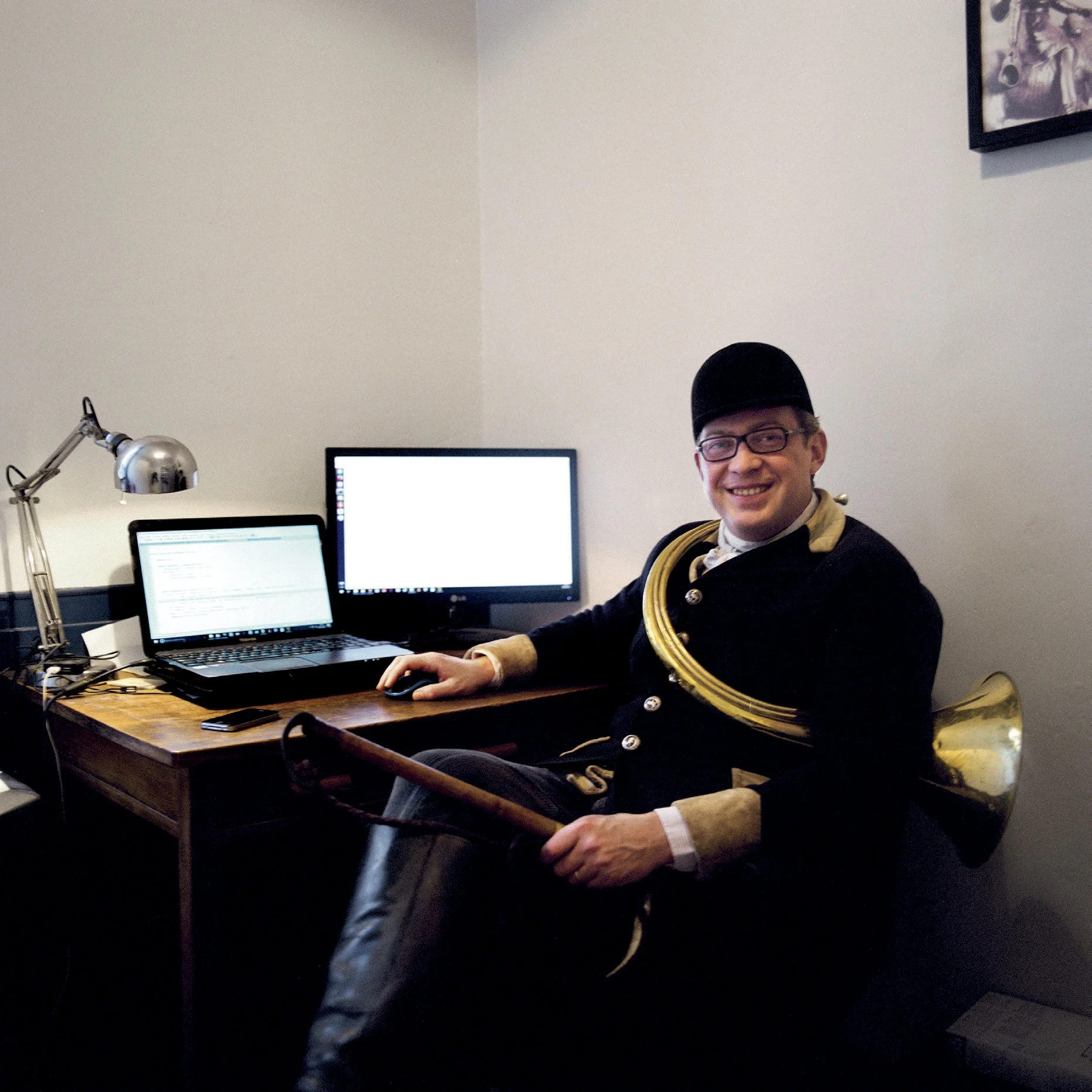 Men in traditional costume and caps, seated at a desk with two computer screens. He smiles at the camera.