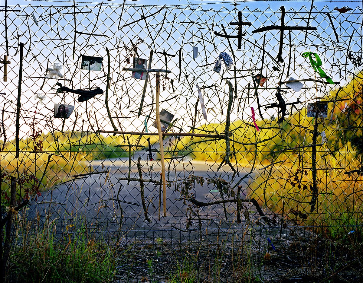 An old wire fence with photos, ribbons and hanging objects, in front of a rural landscape with a clear blue sky.