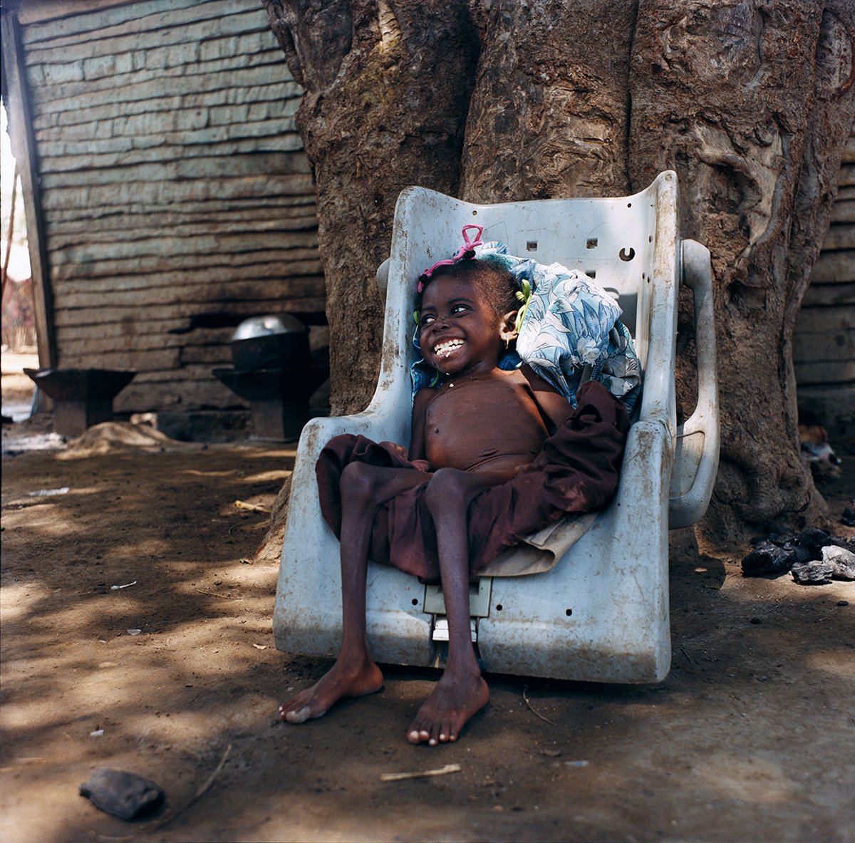 A smiling child sitting in a worn plastic chair under a large tree in a rural setting.