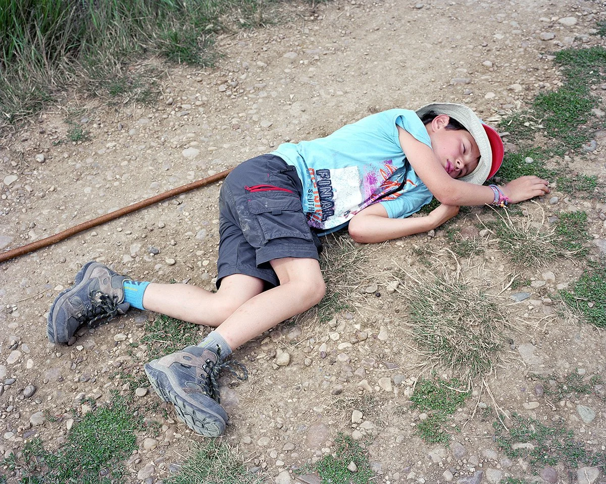 Young boy lying on the dirt floor, baited by fatigue, wearing a hat, blue T-shirt, black shorts and hiking boots, with a branch beside him, in a natural environment.