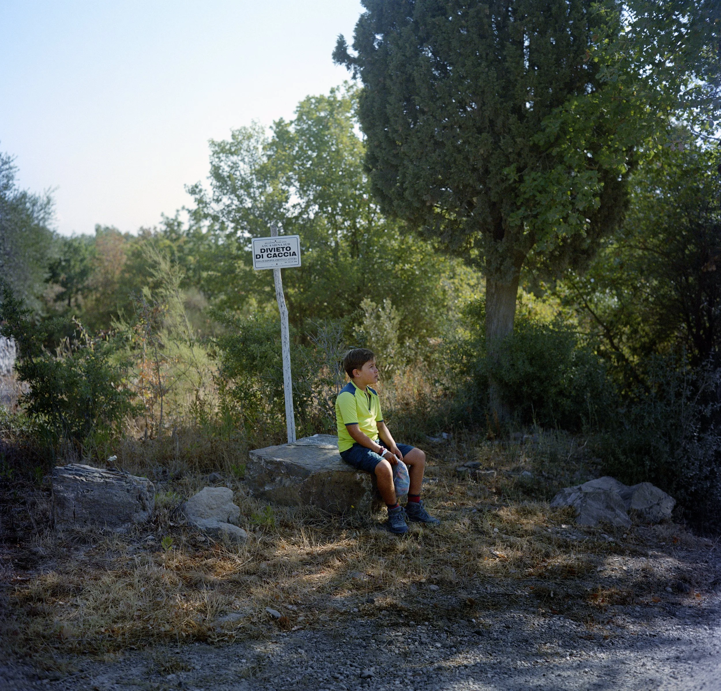 Young boy sitting on a rock in a wooded area, next to a no-hunting sign, with trees in the background.