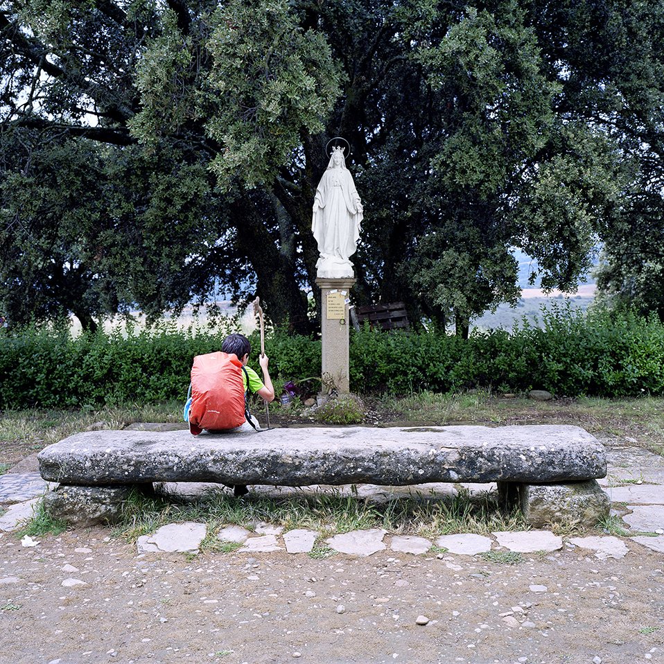 Young boy sitting on a stone bench in front of a statue of the Virgin Mary, surrounded by trees and bushes in a rural setting.