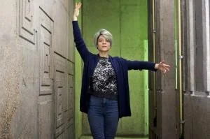 Elderly woman with gray hair, wearing a patterned sweater and blazer, posing with arms raised in an industrial green space.