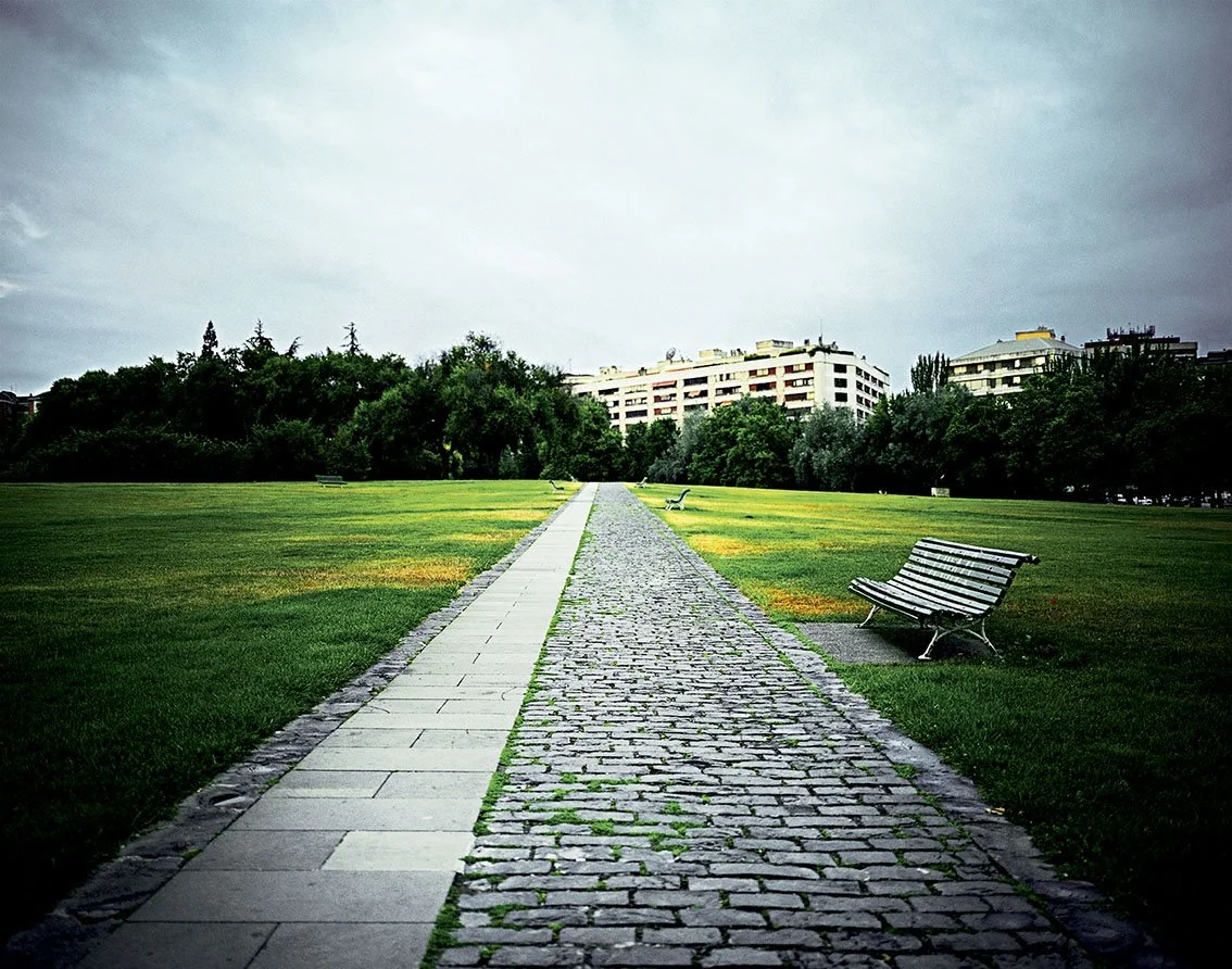 Paved driveway leading to a park with benches, trees and residential buildings in the background, cloudy sky.