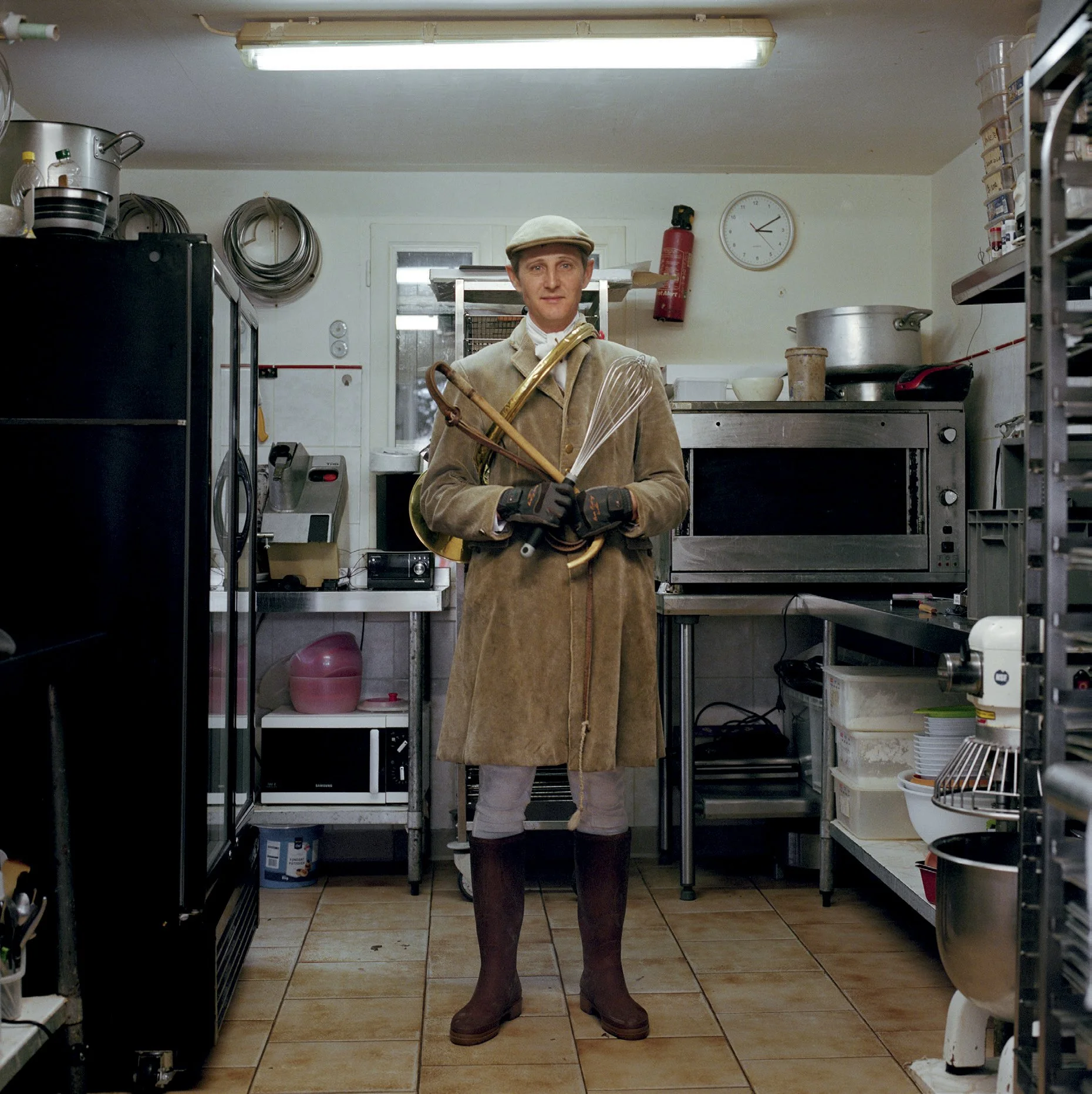 Man dressed in period costume, wearing rubber boots, holding a whip, trapeze and cane in an industrial kitchen.