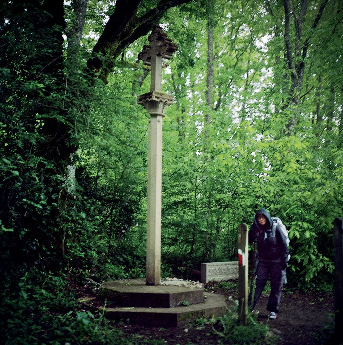 A person hiking through a green forest, passing an old stone calvary with a cross at the top, surrounded by trees and dense vegetation.