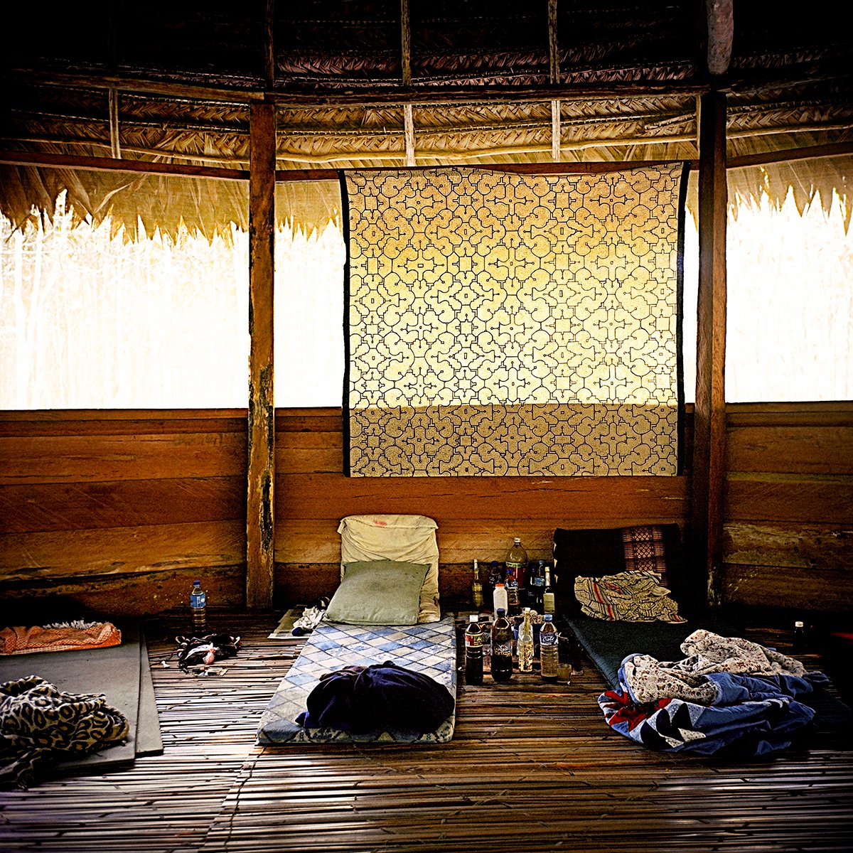 Interior of a wooden hut with a thatched roof, containing mattresses and personal belongings.