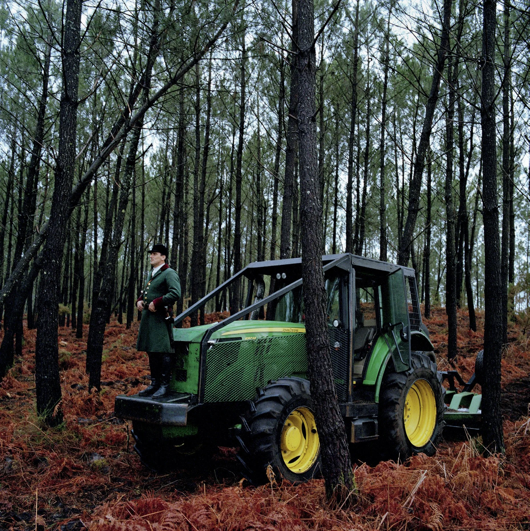 A man in traditional clothing stands on a green tractor in a coniferous forest, with the ground covered in brown ferns.