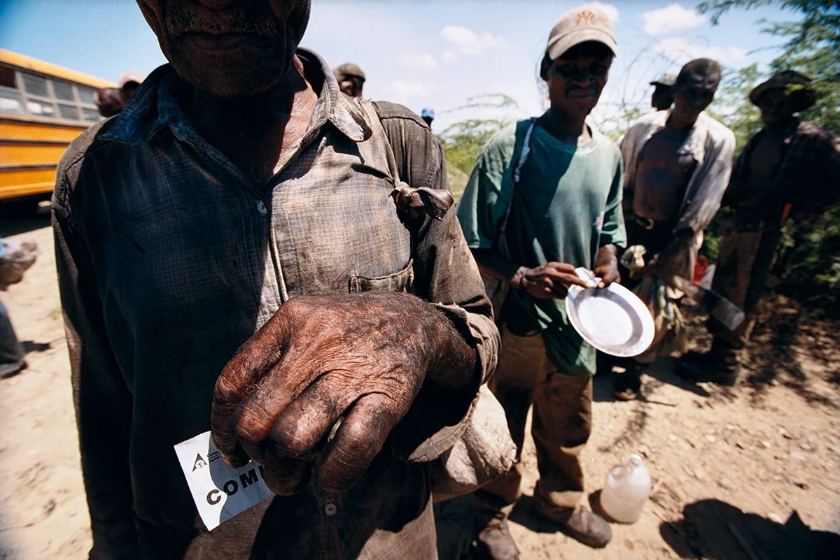 Group of people in an outdoor environment, with a man showing his rough, dirty hand, wearing a badge, other people in the background, a yellow bus visible.
