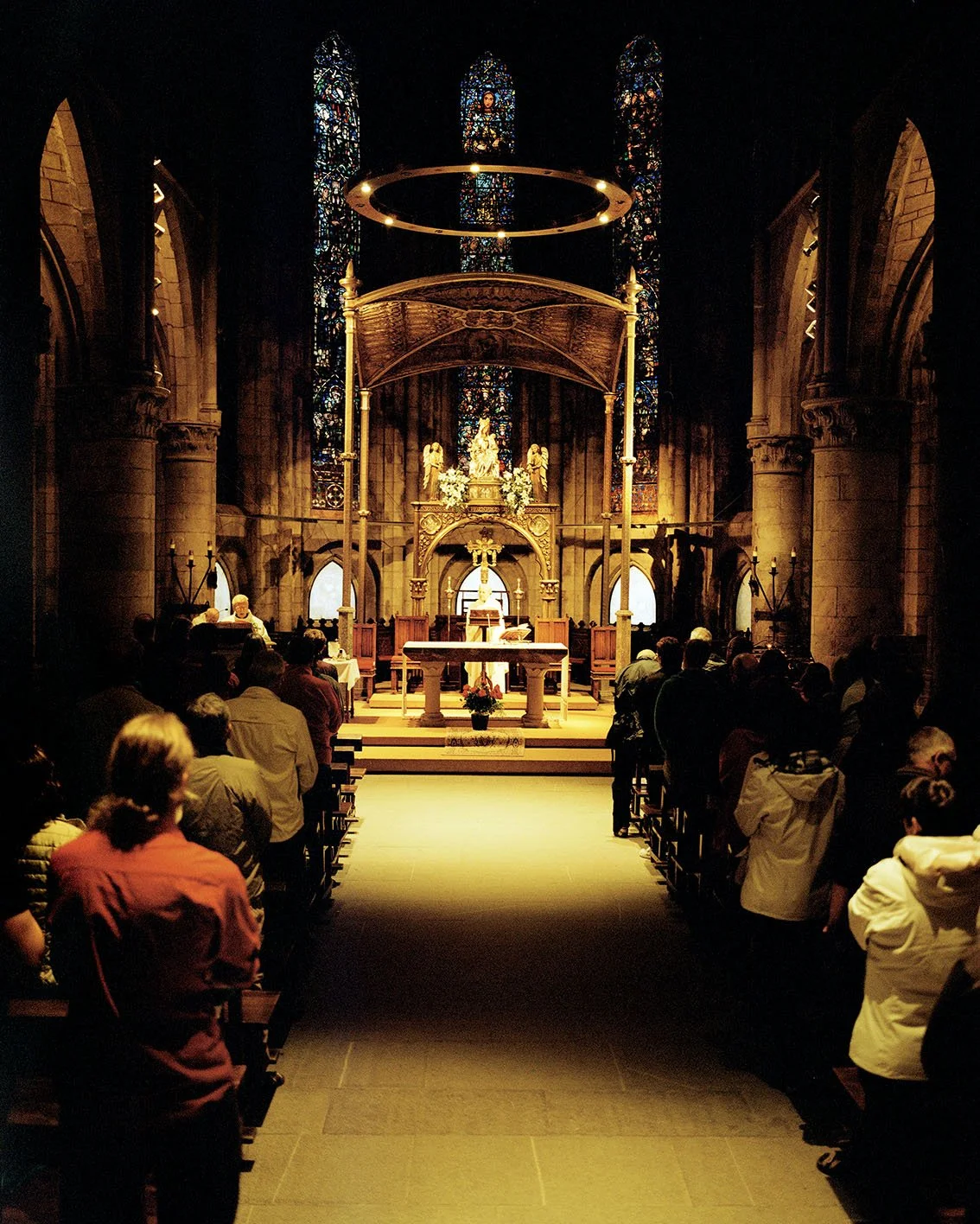 Interior of a Gothic church with a group of people praying or attending a religious service in front of the altar, with colored stained glass behind.