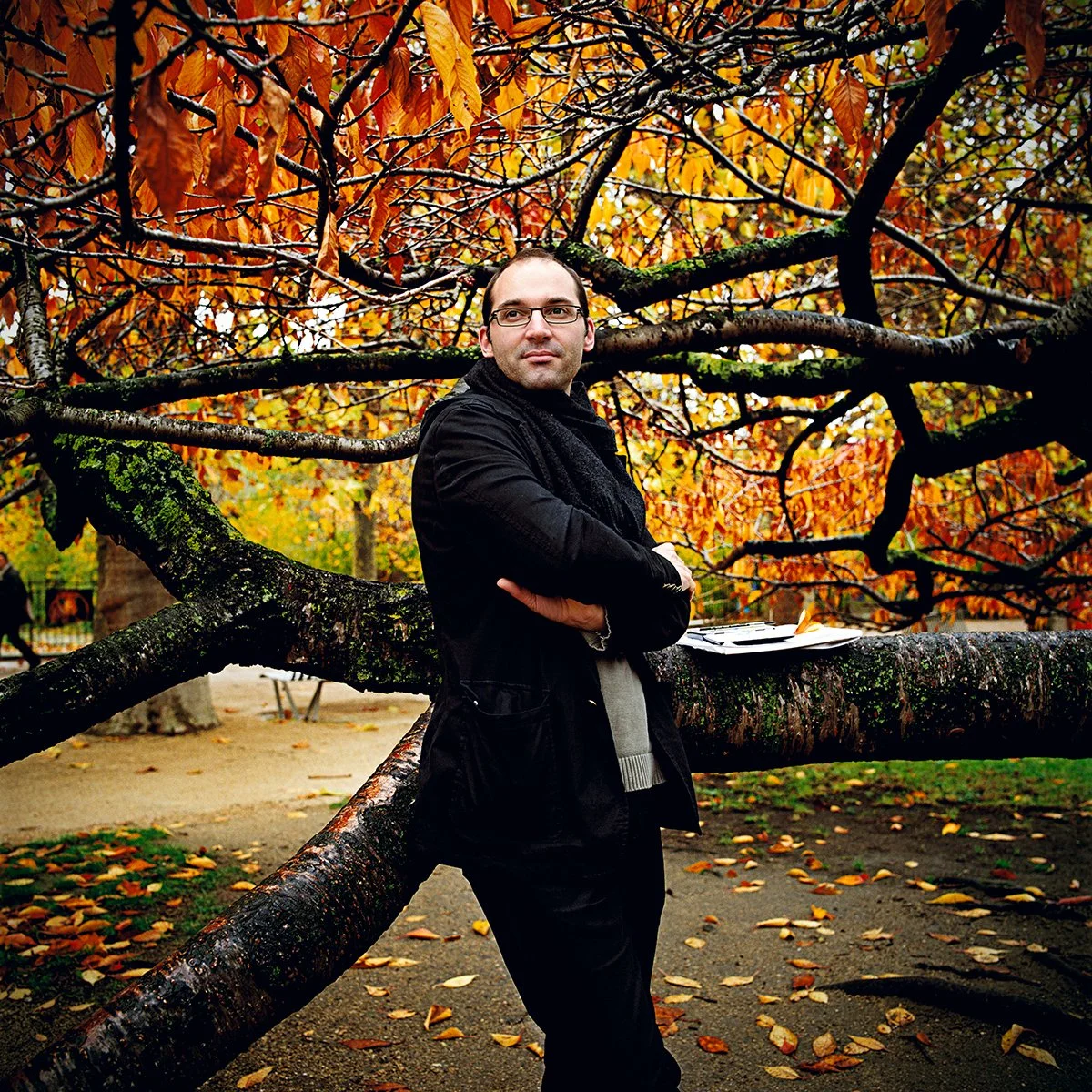 A man standing in front of a tree whose branches are spread out in a park in autumn, with orange and yellow leaves. The man is wearing a black coat and glasses, and stands with his arms crossed.