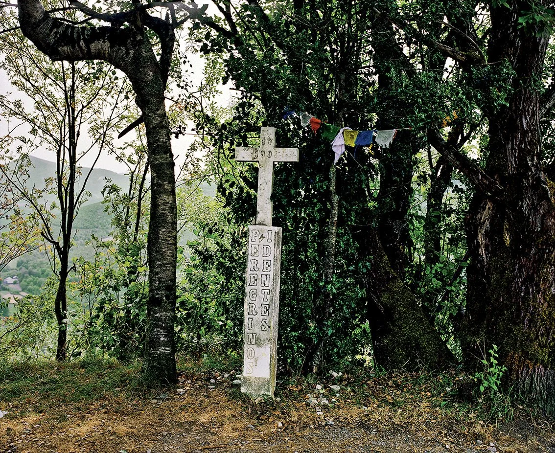 Stone cross with inscription, set between two trees, with colored prayer ribbons hanging from it, in a forest setting.