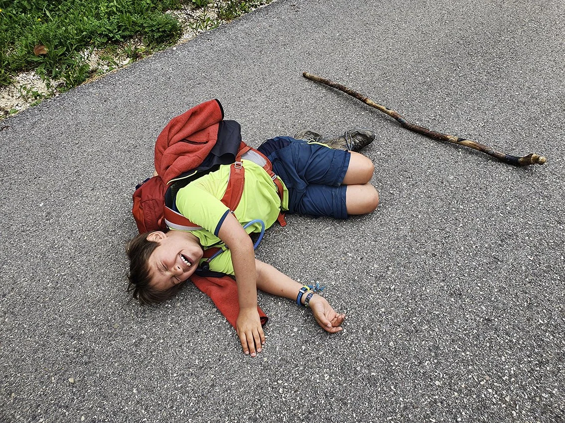 A young person lying on the ground on a road with a smile on his face, wearing a backpack, a green T-shirt and blue shorts, with a stick by his side.