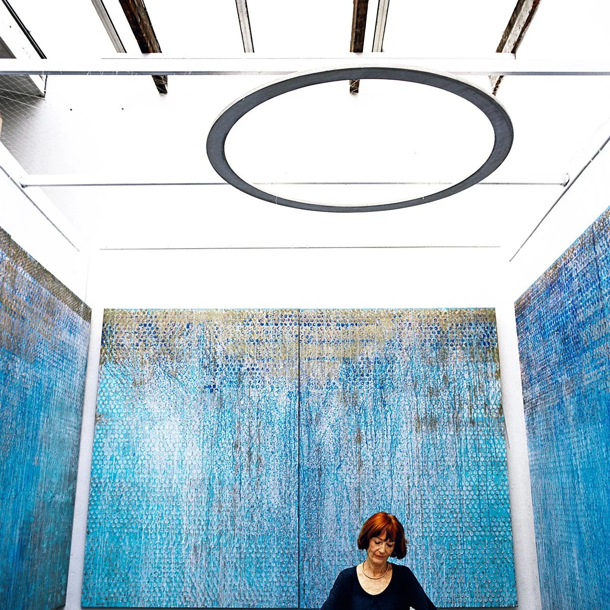 A woman with red hair looks down into an exhibition room with blue abstract art hanging on the walls behind her. A large black circle hangs from the ceiling.