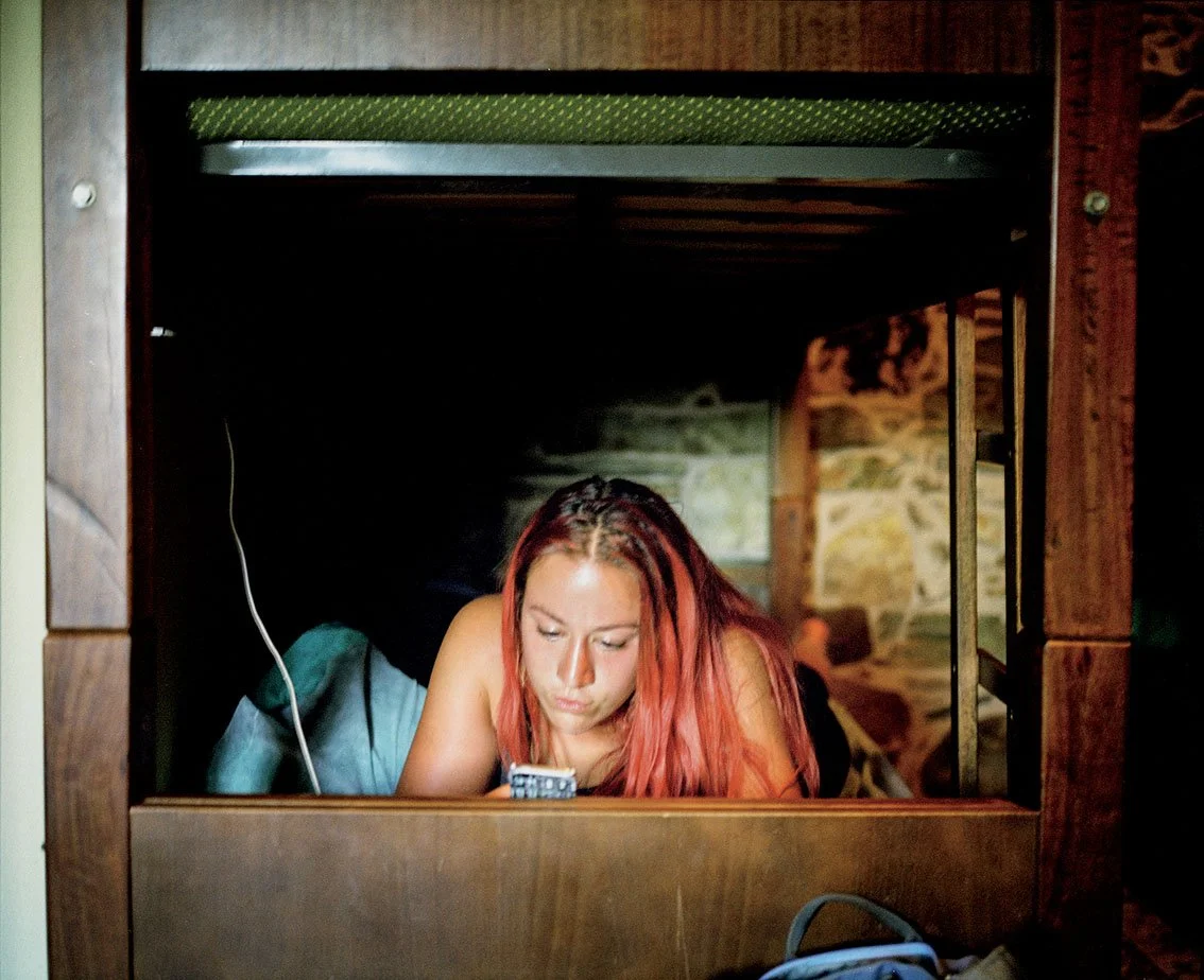 Young woman with red hair, lying under a wooden desk, staring at her cell phone.