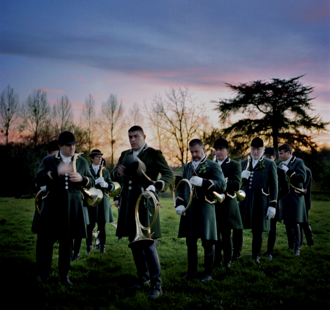 Un groupe de musiciens en uniformes traditionnels, tenant des cors en métal, dehors au crépuscule. Arbres sans feuilles en arrière-plan, ciel coloré de rose et de bleu.