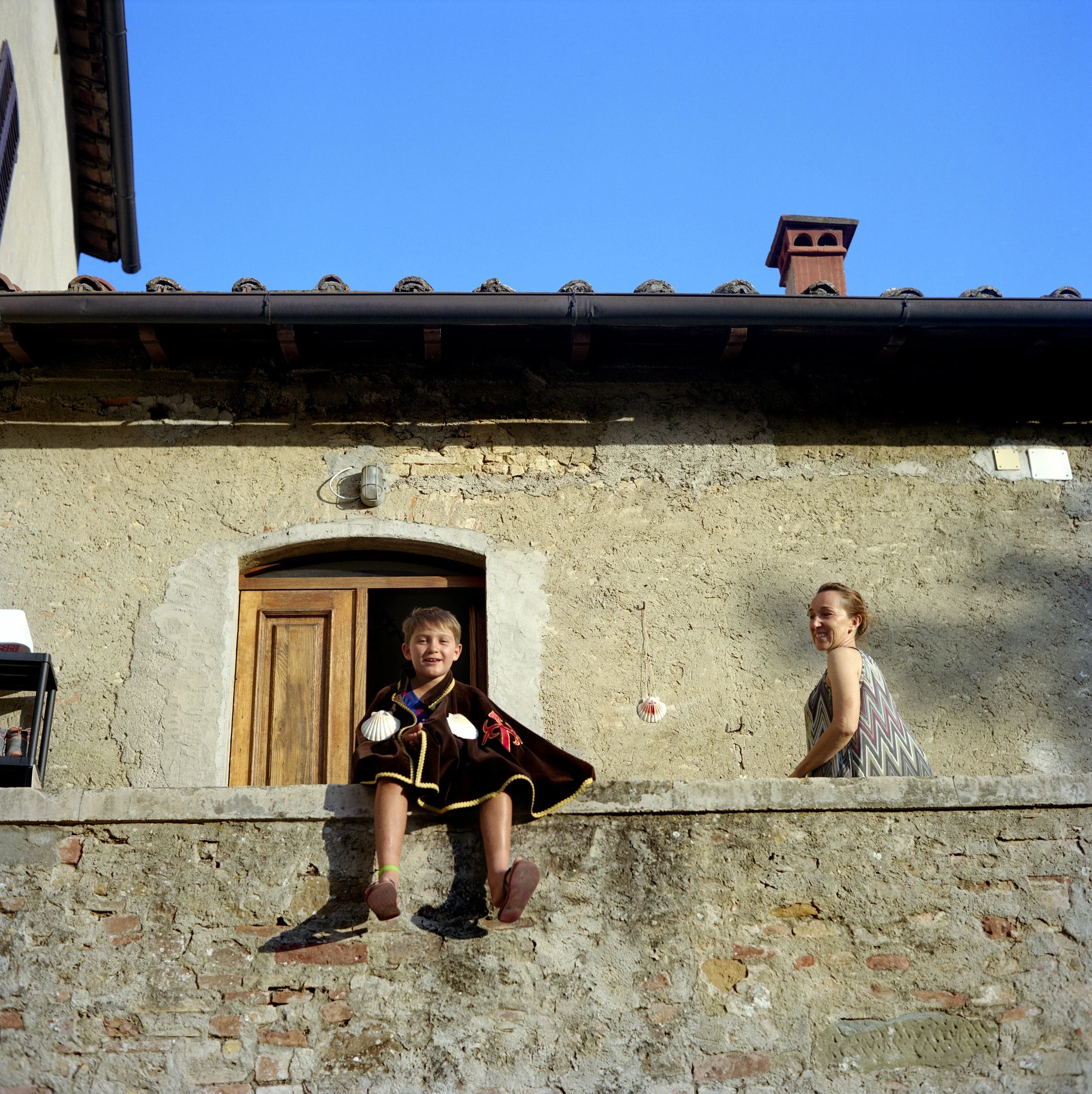 A young boy dressed like a hero, sitting on the wall of a stone house with a woman next to him, both smiling, under a blue sky.