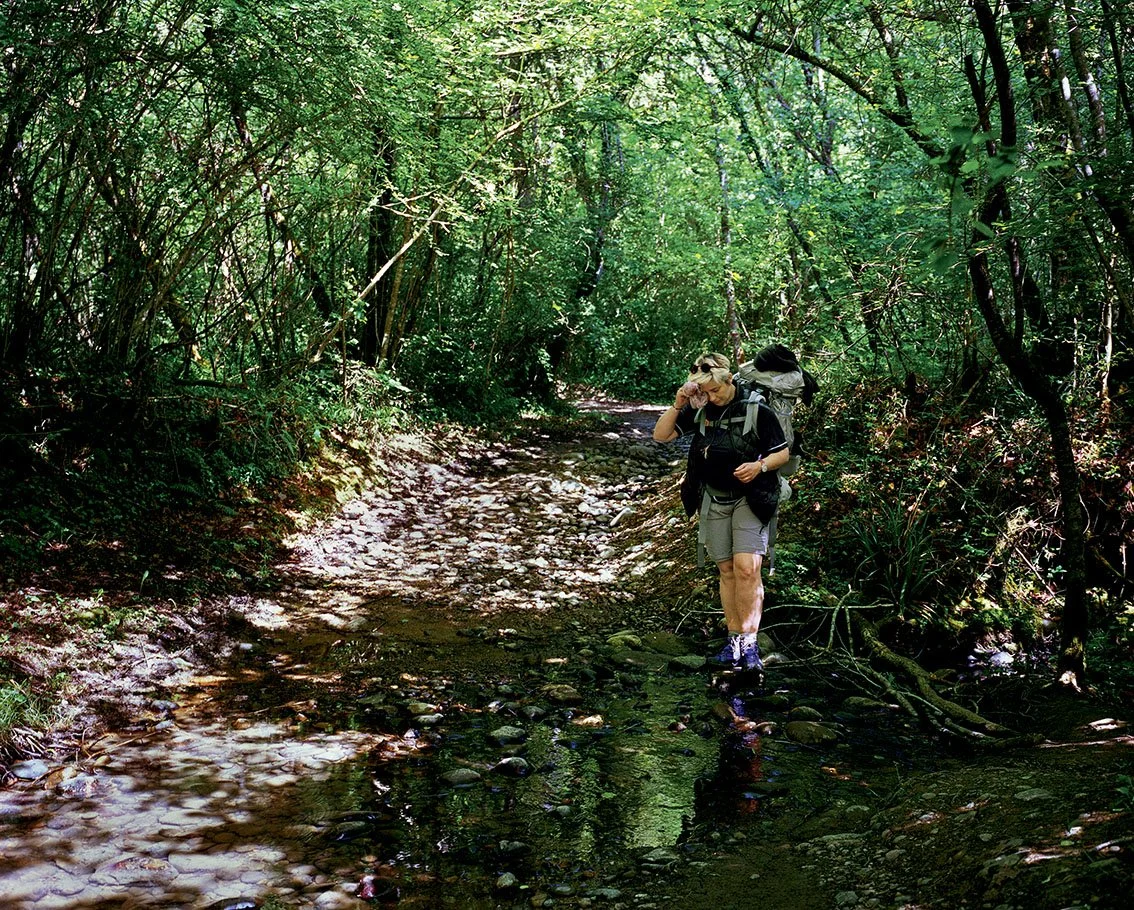 A woman walks along a forest path carrying a backpack, surrounded by dense vegetation and green foliage.