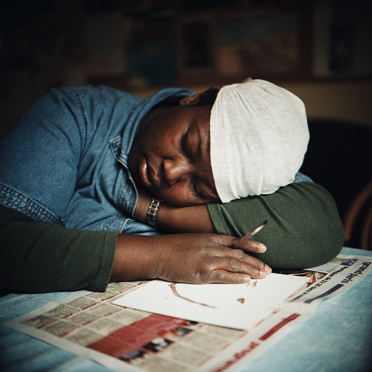 Une femme repose sa tête sur ses bras, portant un bonnet blanc, assise à une table avec des journaux, semblant dormir ou être épuisée.