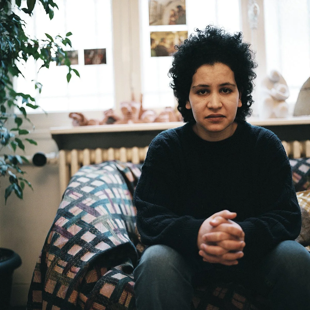 Une jeune femme aux cheveux bouclés assise sur un canapé, regardant l'objectif, dans une pièce lumineuse avec des fenêtres et des décorations en arrière-plan.
