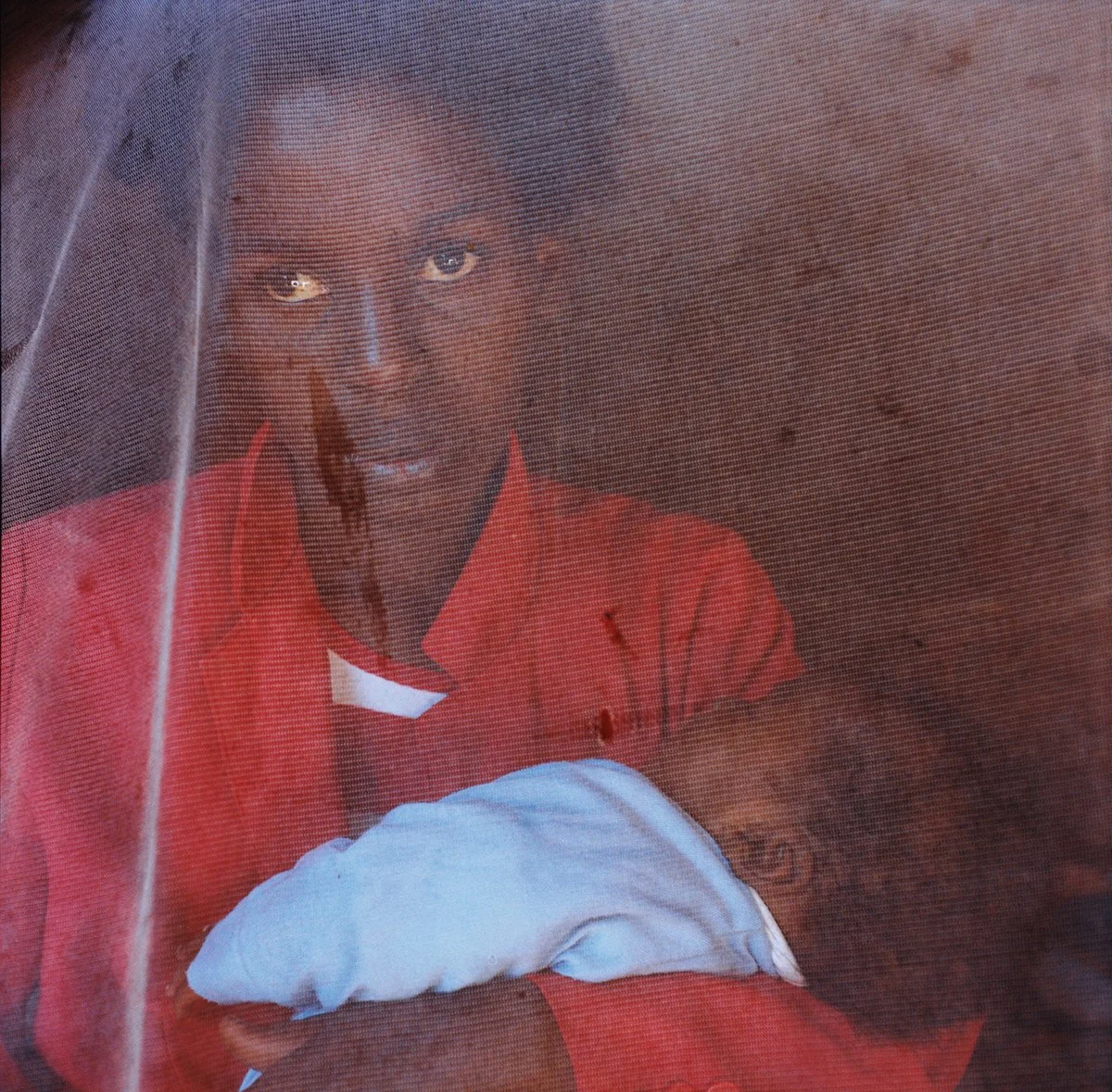 A person behind a net or pierced canvas, wearing red clothing, with a dark face and serious expression.