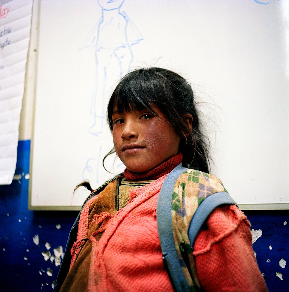 Young girl with a backpack, wearing a red sweater, in a classroom with a whiteboard and a drawing of a girl in the background.