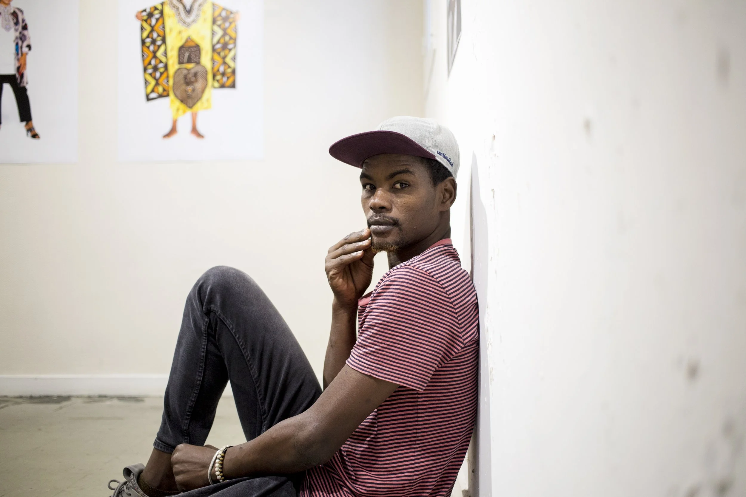 Young man sitting against a white wall in an art gallery, wearing a striped t-shirt and cap, looking towards the lens.