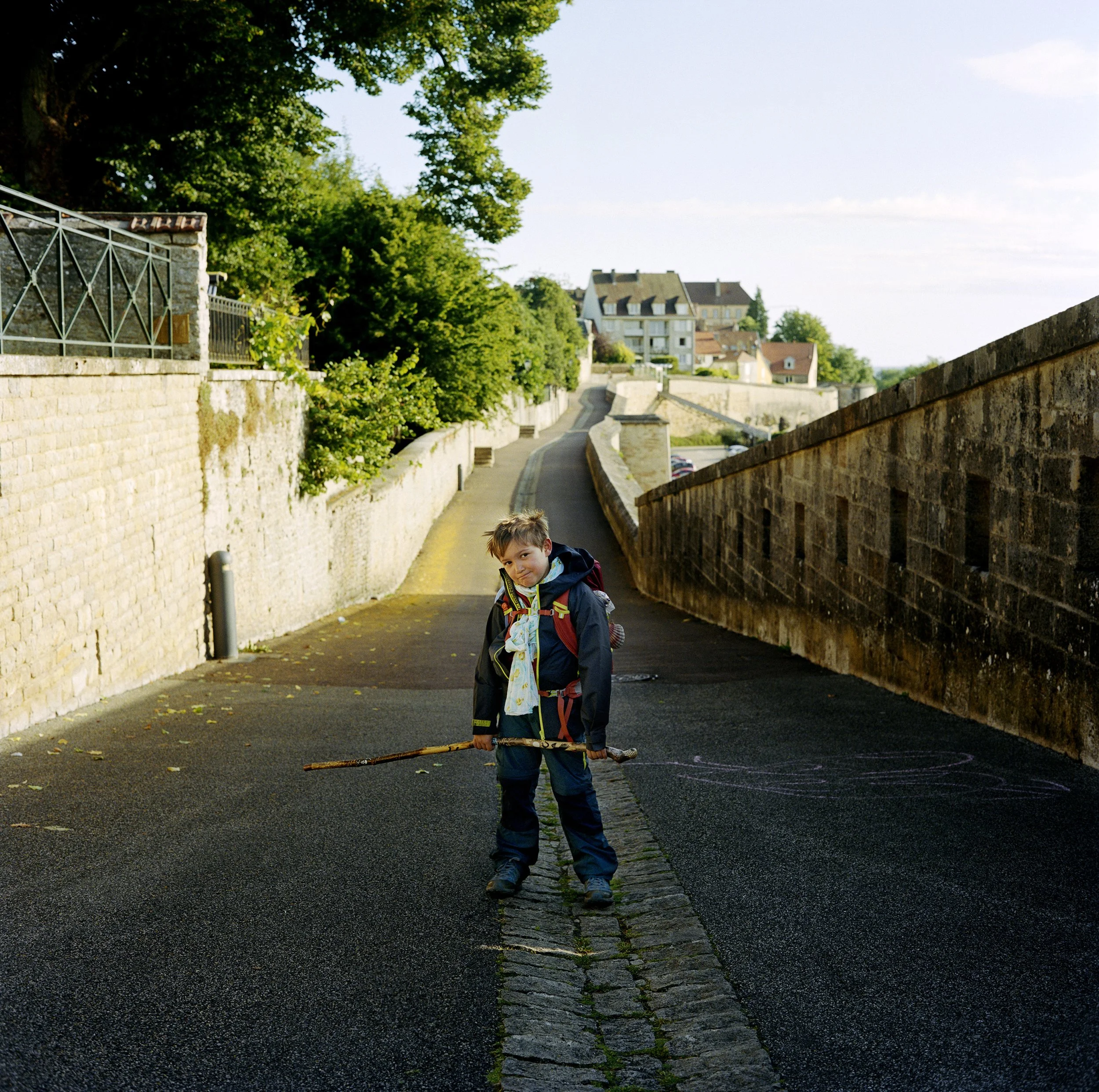 Un jeune garçon avec un sac à dos et un bâton dans une rue en pente avec des murs en pierre, des maisons en arrière-plan et des arbres verts.