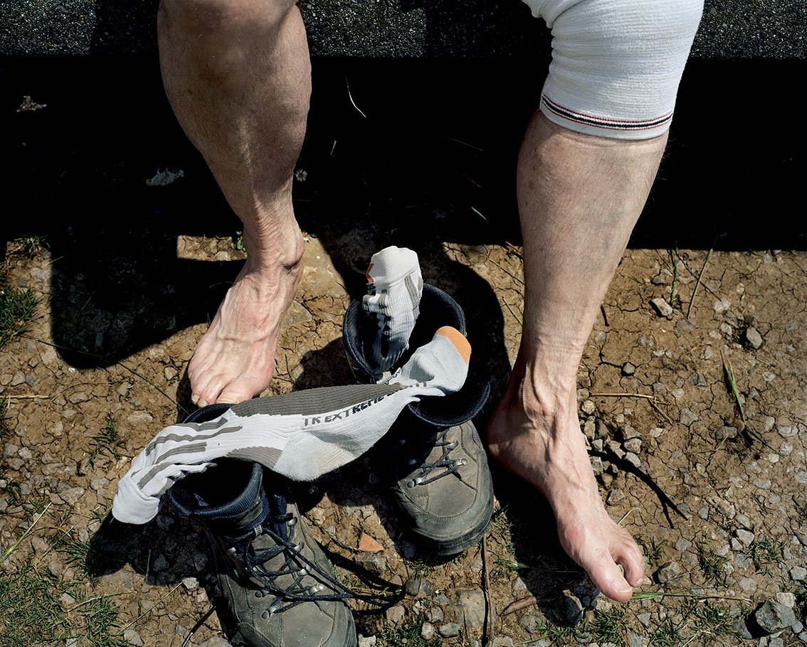 Bare feet of a person, wearing a sock and walking shoes, in front of sports socks on boots, on a dry earth floor.