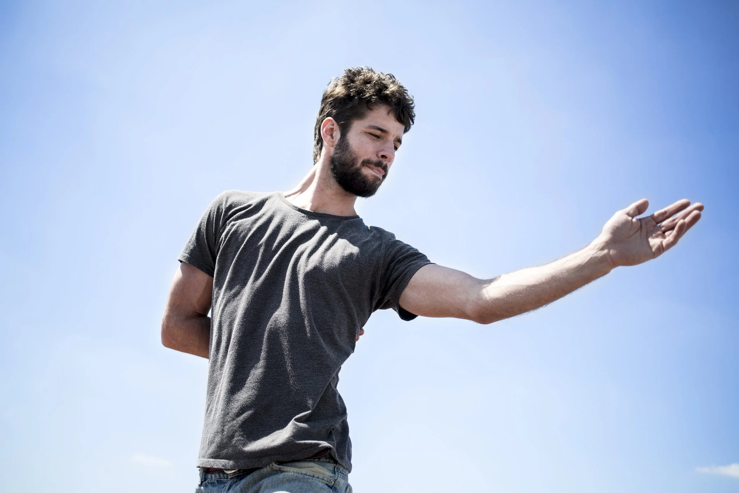 Man with beard, wearing a grey T-shirt, arm stretched forward, under a clear blue sky