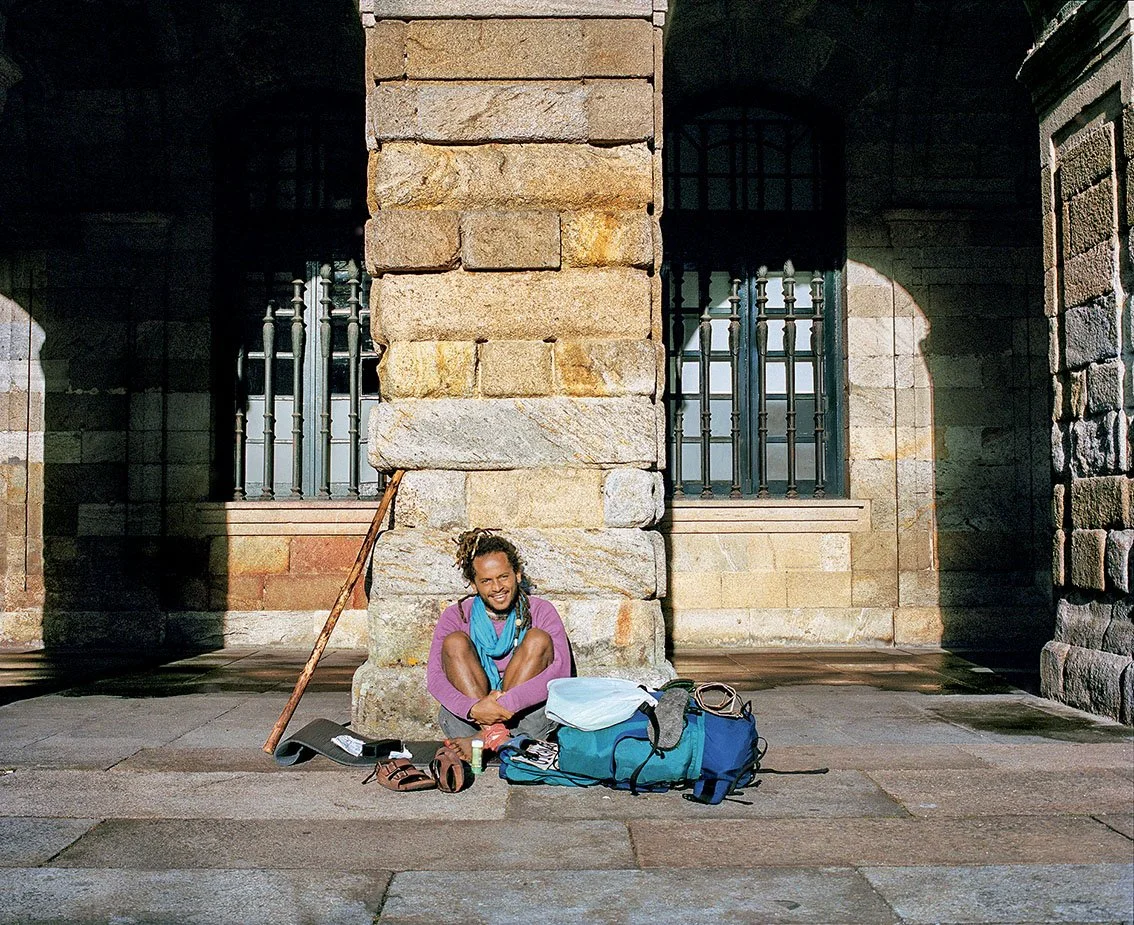 A person sitting on the ground in front of a stone building, with a backpack, hiking boots, walking stick and other objects, smiling, in broad daylight.