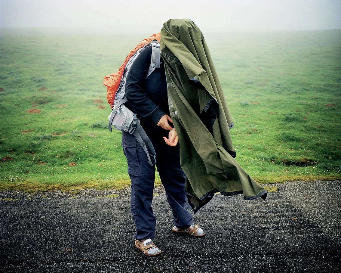 A person in hiking clothes holding a green raincoat about to put it on, in a misty landscape with green grass and black soil.