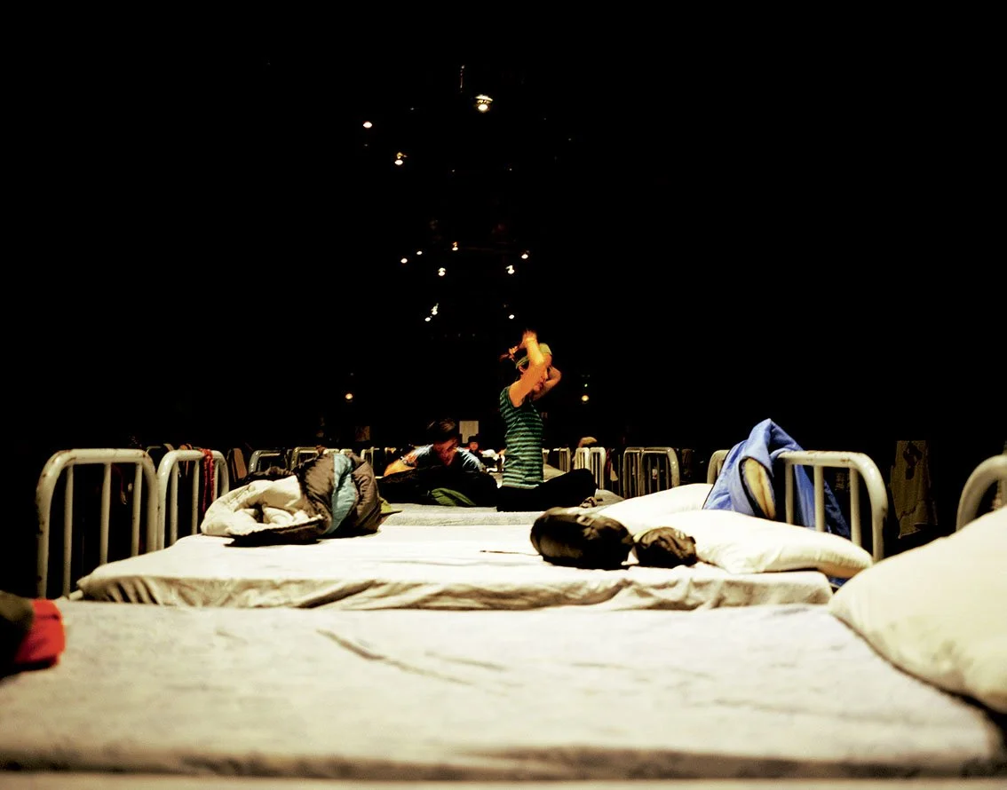 Beds or mattresses with luggage and one person sitting in the middle in a dark room.