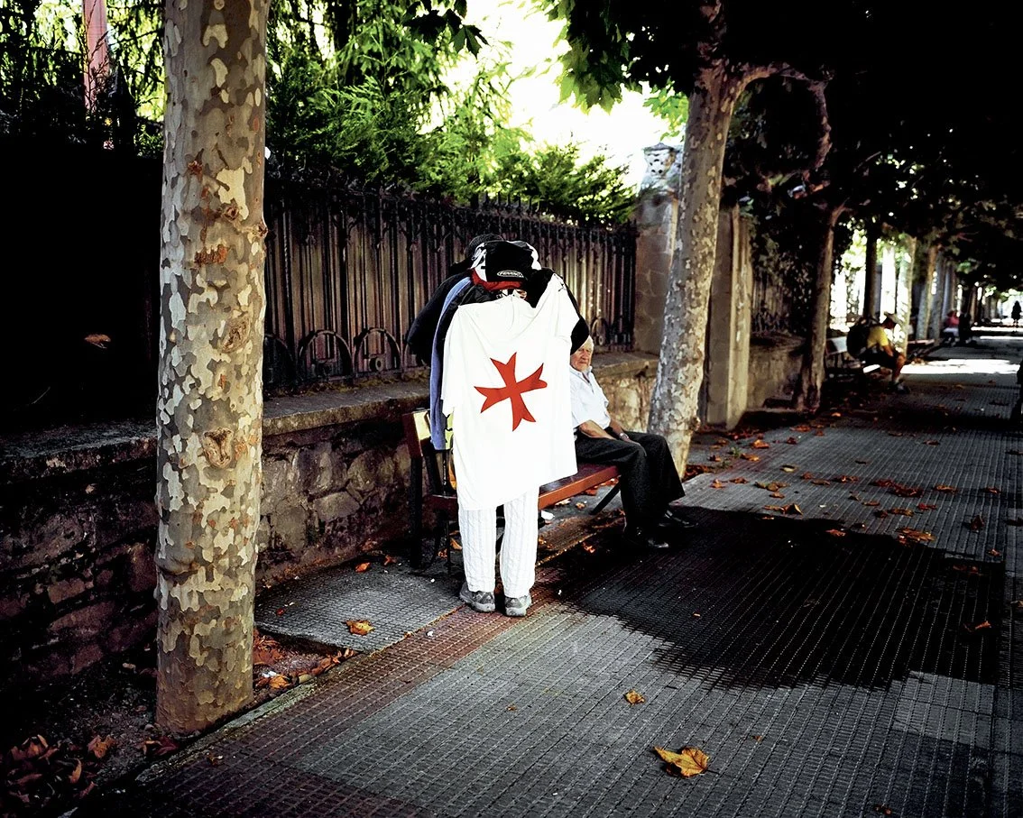 A person with an Order of Malta flag stands in front of someone lying on a bench in a street lined with trees and seated people.