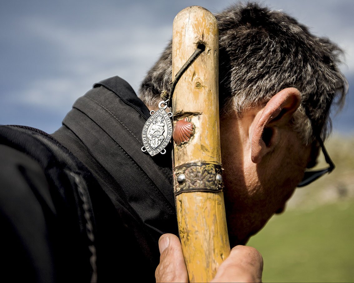A man carries a long stick on his shoulder and rests his ear against it, in an outdoor landscape with a cloudy sky.