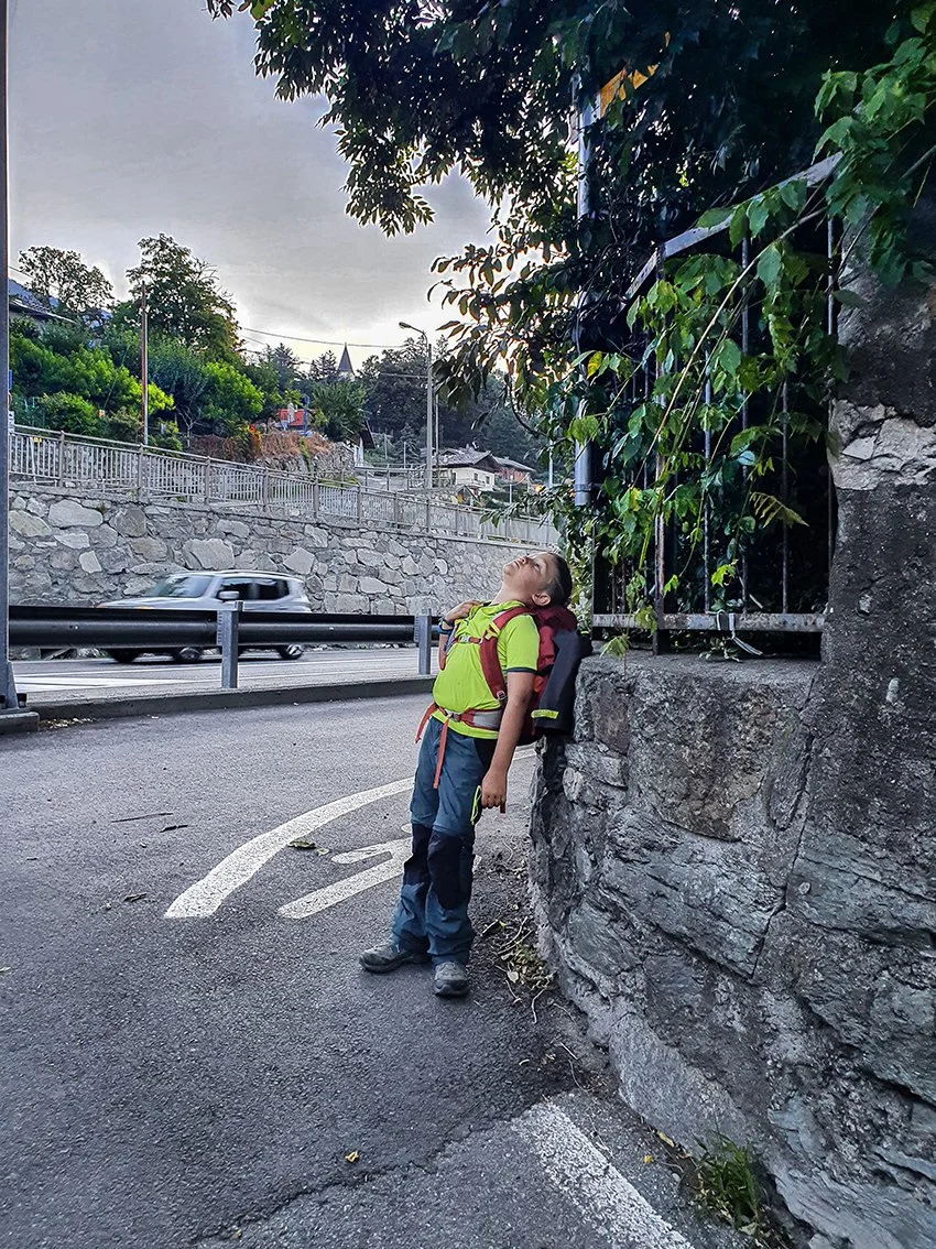 A child with a green and red backpack, dressed in hiking gear, stands on the side of the road, leaning against a rock face, looking up carelessly.