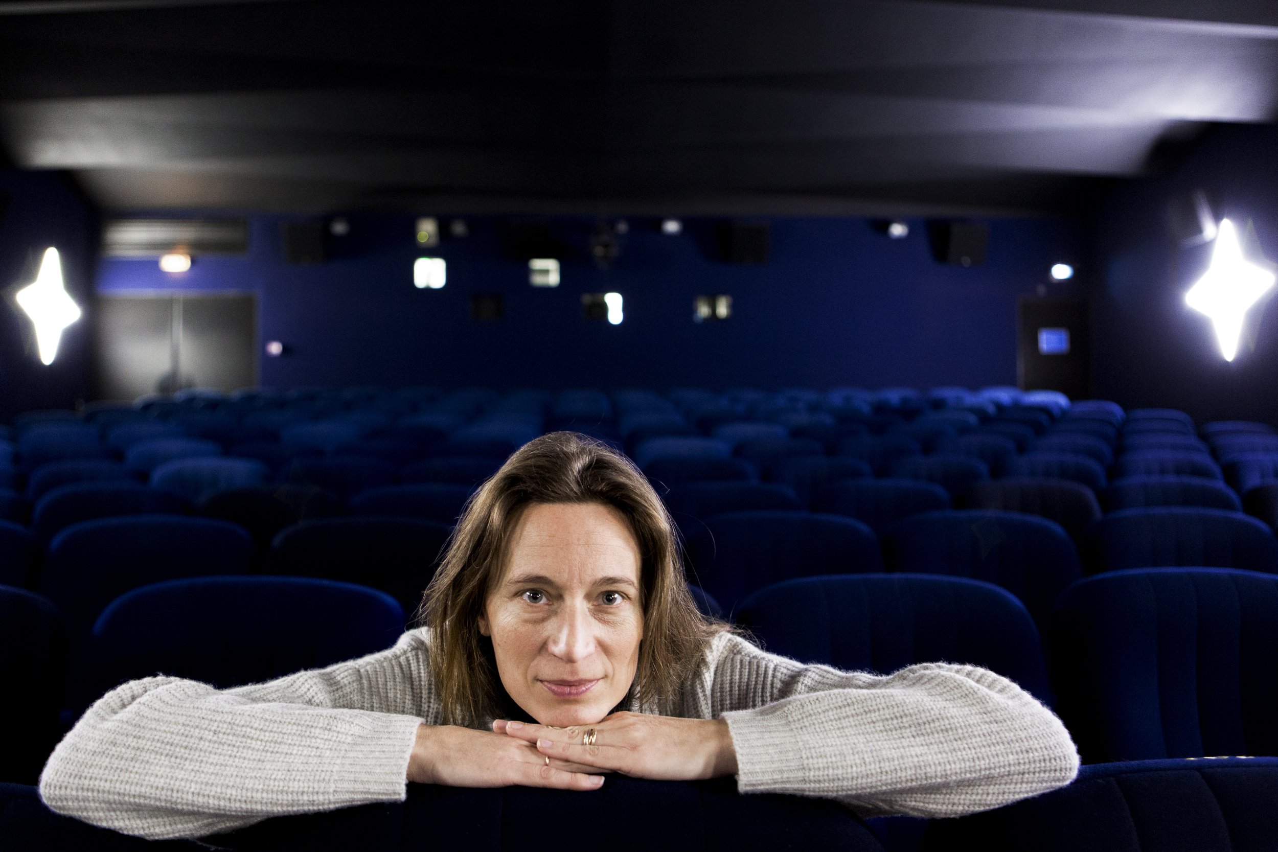 A woman sitting in an empty cinema, leaning on her arms, with dim lighting and blue seats.