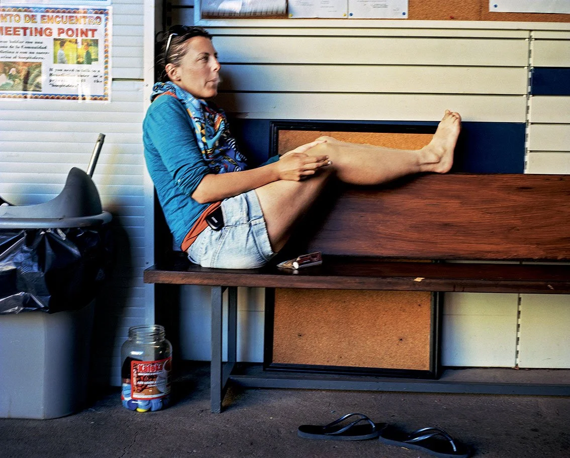 A woman sitting on a bench with one leg outstretched, wearing a blue T-shirt, blue bandana, and shorts, in a room with a paneled wall and various objects around it.