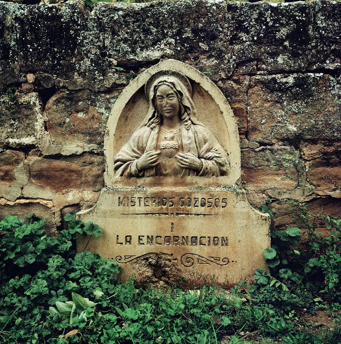 A sculpture of Mary in a rock niche with the plaque inscribed "Mystérios Gozosos à La Encarnacion", surrounded by greenery.