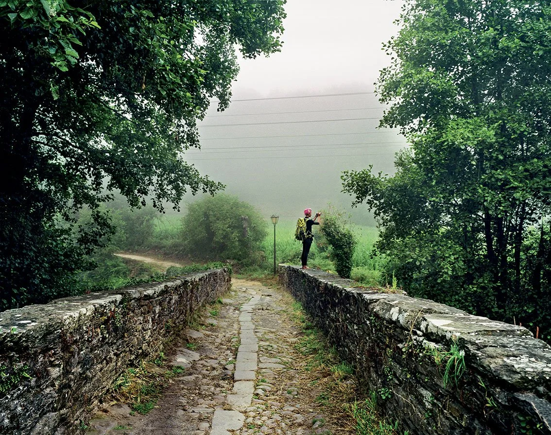 A person wearing a pink helmet photographs a misty landscape on an old stone bridge surrounded by green trees.