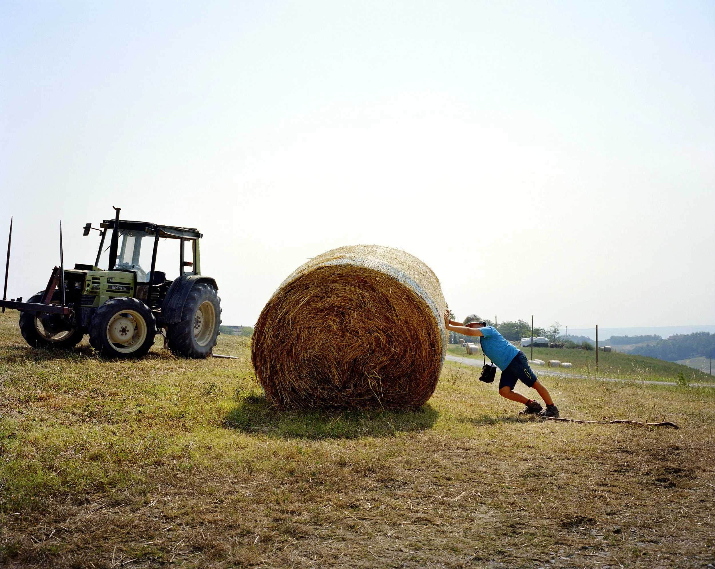 A child pushes against a large bale of hay in a field with a tractor in the background, clear sky.
