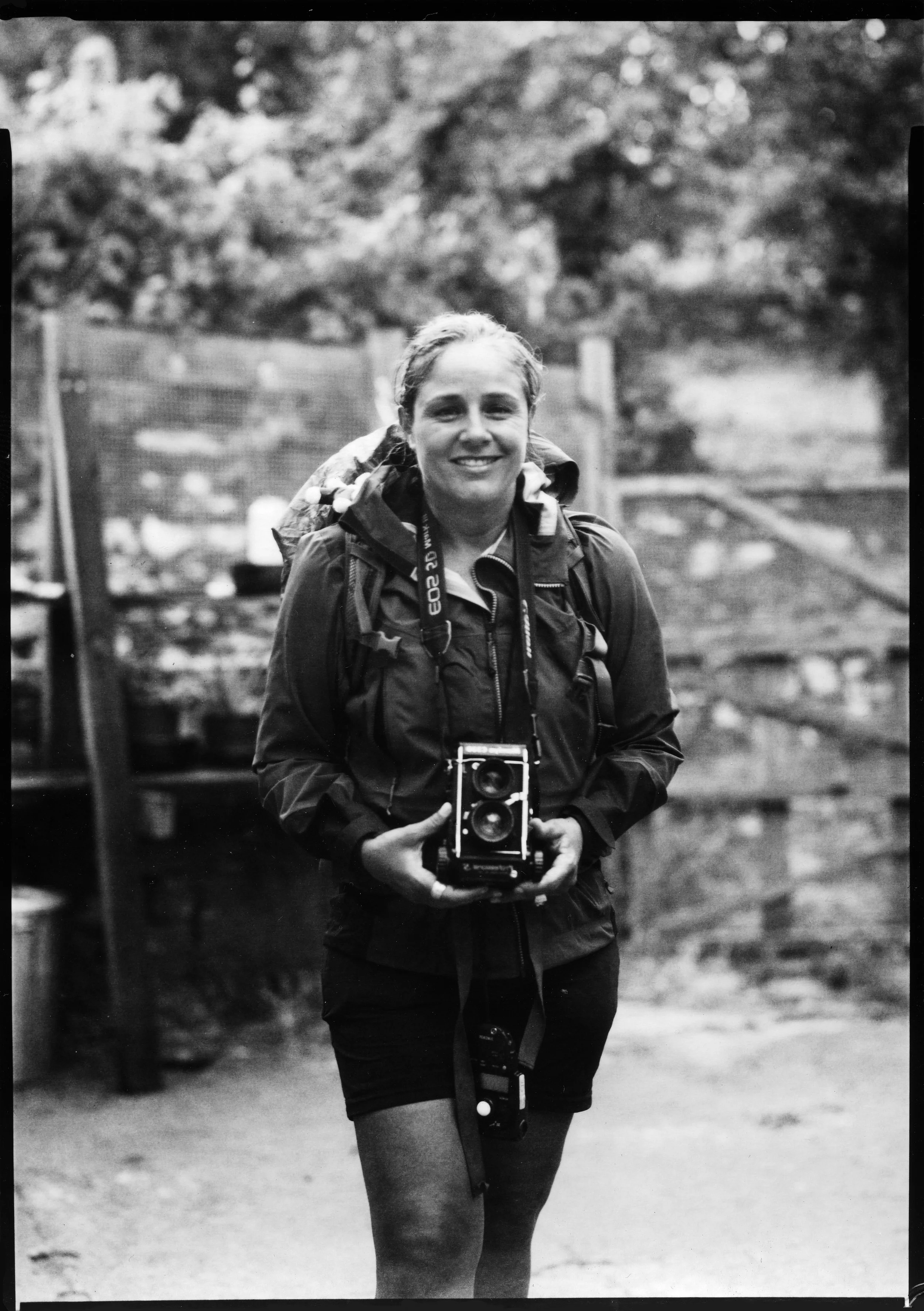 Une femme souriante en randonnée portant un sac à dos et tenant un appareil photo, en extérieur.