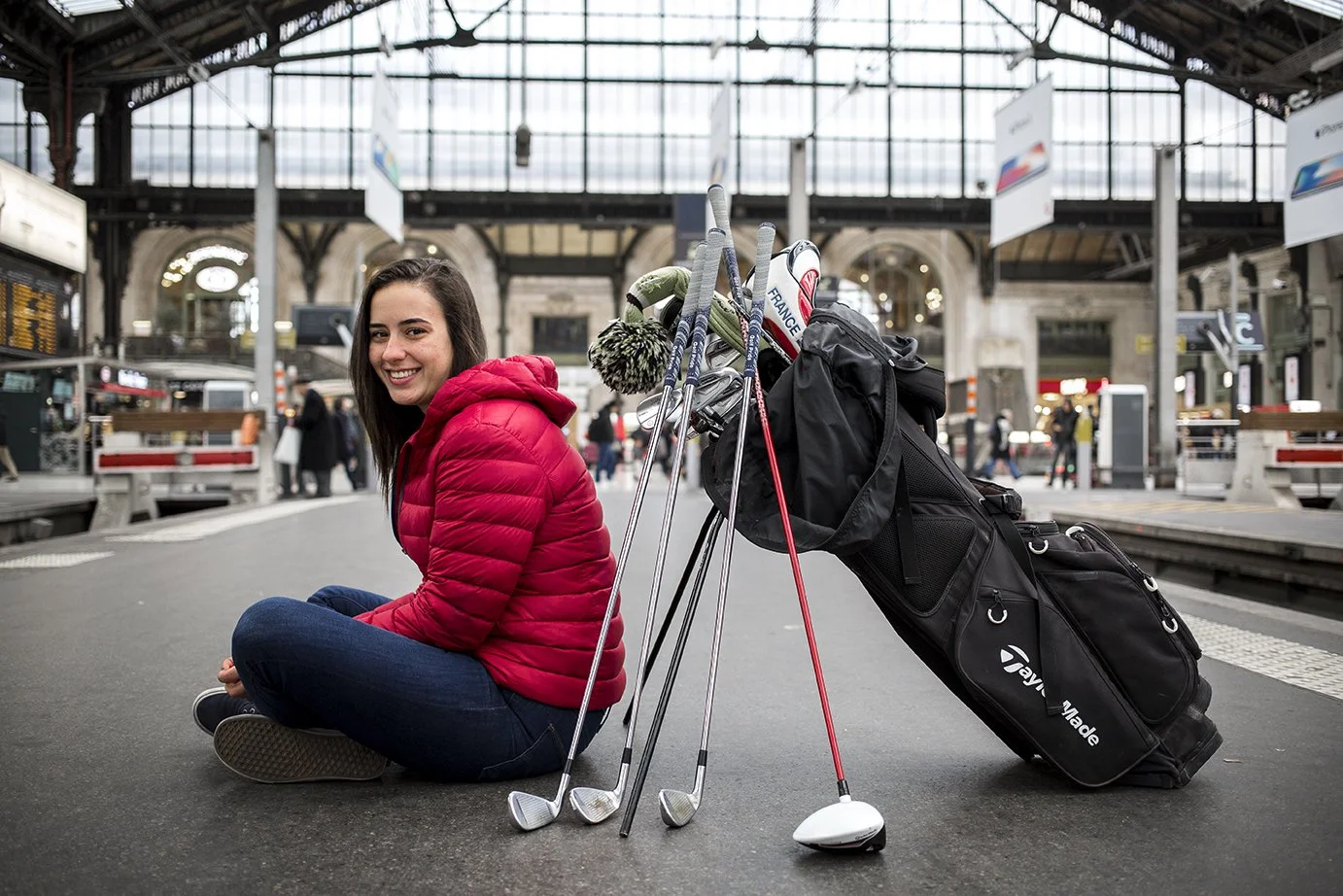 Smiling young woman sitting on the floor of a train station with a golf bag and clubs, wearing a red jacket.