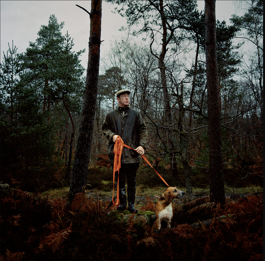 Un homme avec un chapeau et un manteau noir tient une laisse orange attachée à un chien de race beagle, dans une forêt en automne avec des arbres nus et verts autour.