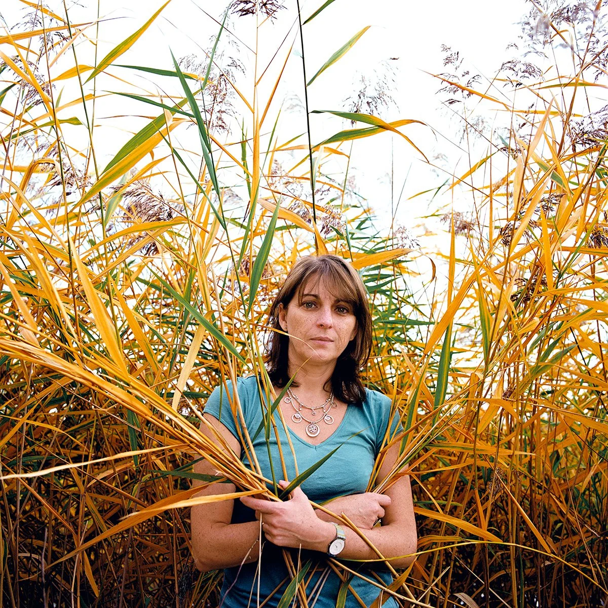 Woman standing in a field of golden reeds, wearing a blue T-shirt and silver necklace.