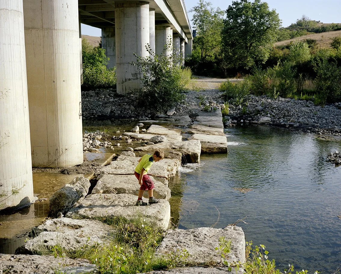 A boy is playing near a river under a concrete bridge with large pillars. He is walking on rocks at the water's edge, surrounded by green vegetation.